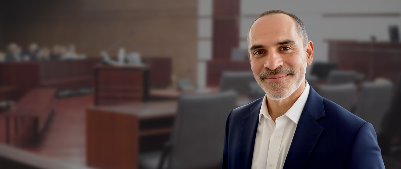 Portrait of Joe Serpa, dressed in a professional blue suit with a patterned tie, standing in the foreground with a blurred courtroom background.