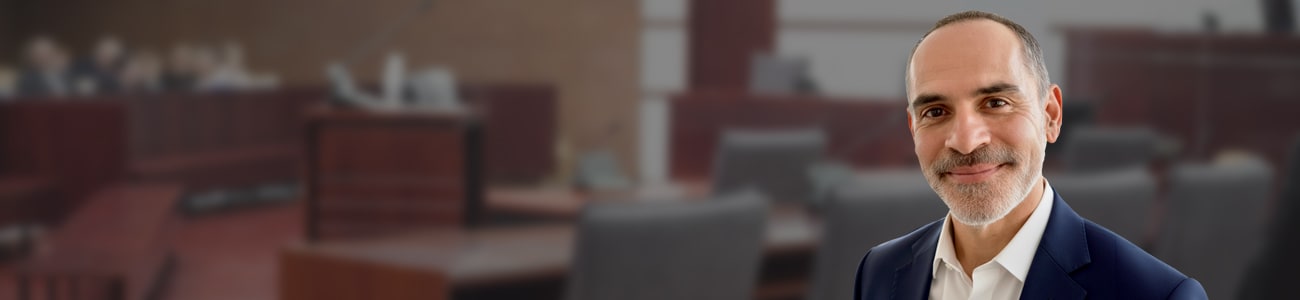 Portrait of Joe Serpa, dressed in a professional blue suit with a patterned tie, standing in the foreground with a blurred courtroom background.