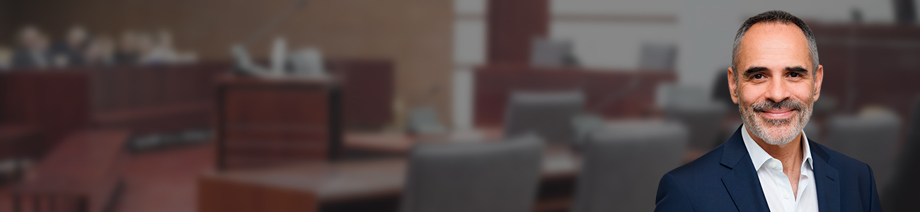 Portrait of Joe Serpa, dressed in a professional suit, standing in the foreground with a blurred courtroom background.